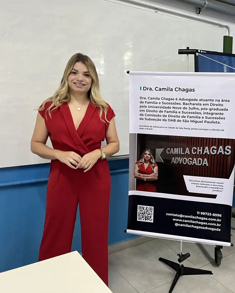 Dra. Camila Chagas, de macacão vermelho, ao lado de um banner com o nome e a apresentação dela, numa sala de aula